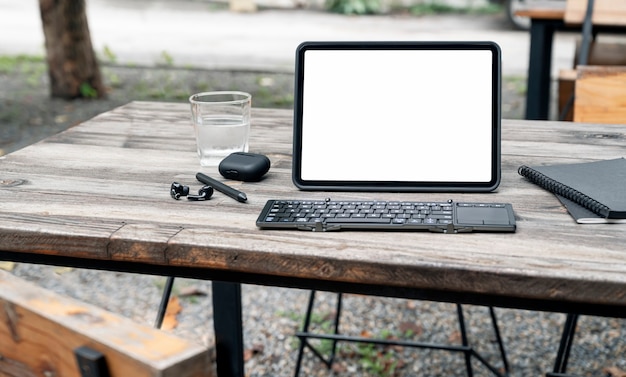 Makieta pustego ekranu tablet, klawiatura, słuchawki, rysik i szklankę wody na drewnianym stole.