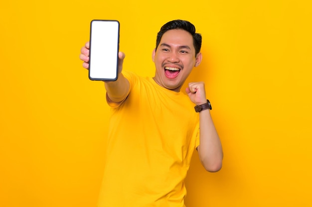 Makieta pustego ekranu telefon podekscytowany młody człowiek azjatyckich w casual tshirt pokazano telefon komórkowy z pustego ekranu, co zwycięzca gest na białym tle na żółtym tle koncepcja życia ludzi