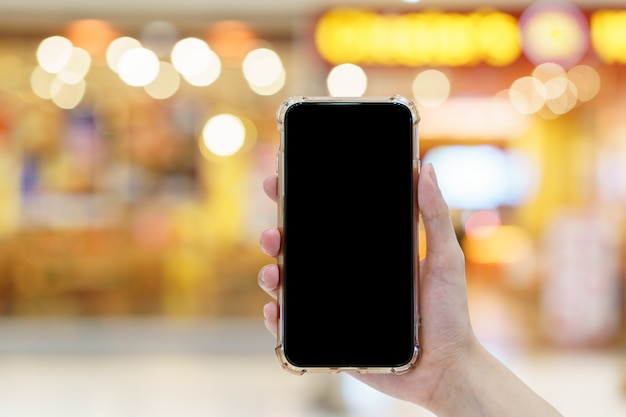 Makieta, ręce trzymając pusty czarny ekran telefon komórkowy w niewyraźnym domu towarowym, koncepcja płatności cyfrowej
