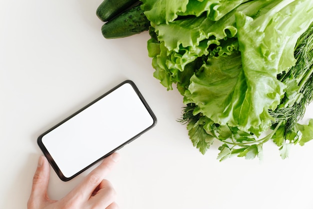 Makieta smartfona i eko warzywa widok z góry Ręka dotykająca telefonu komórkowego z pustym białym ekranem i warzywami organicznymi w ogrodzie na białym stole zbliżenie Zdrowa żywność i miejsce na reklamę