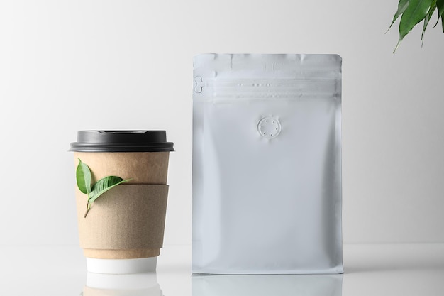 Makieta torebki kawy i filiżanki kawy na szarym tle z zielonymi liśćmi. Kawowa szara torebka zapinana na zamek.