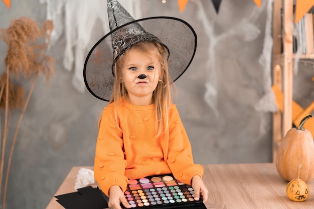 Makijaż na Halloween dziewczynka w pokoju na stole z paletą cieni w dłoniach przygotowuje się do wakacji Pocieranie dziecka w kapeluszu czarownicy Koncepcja wakacji w domu Halloween