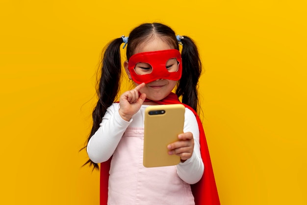 Mała azjatycka dziewczynka w kostiumie supermana i masce używa smartfona koreańskiego dziecka w pelerynie superbohatera