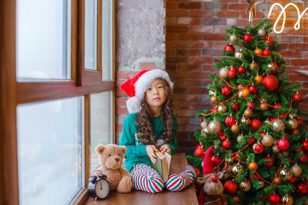 Mała Azjatycka dziewczynka w piżamie siedzi na oknie w pobliżu choinki z niedźwiedziem Boże Narodzenie nowy rok