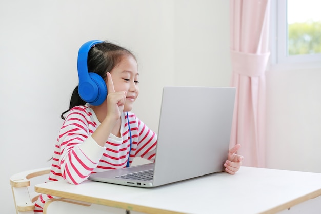 Mała azjatycka uczennica za pomocą słuchawek uczy się lekcji online przez połączenie wideo na laptopie