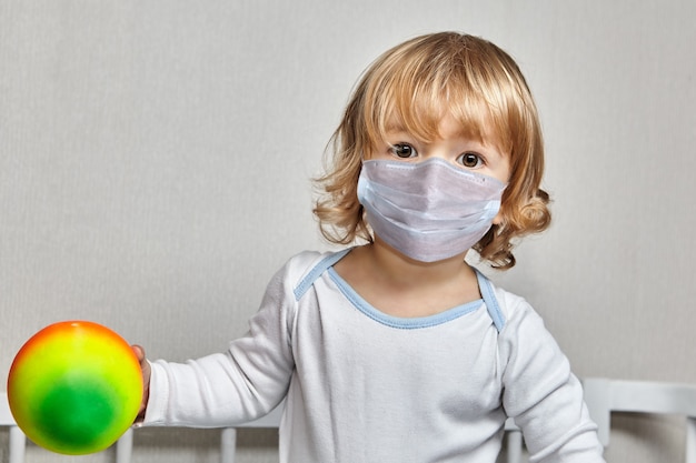 Mała biała dziewczynka w wieku około 3 lat w masce na twarzy bawi się piłką w izolacji domowej podczas pandemii COVID-19.