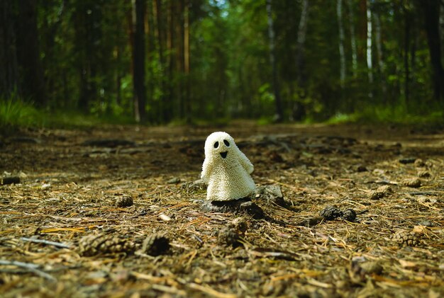 Mała biała zabawka ducha stoi na ziemi w lesie wśród starych igieł i szyszek