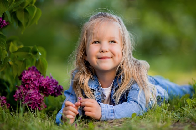 Mała blondynka leży w pobliżu kwiatów bzu na zielonej trawie i uśmiecha się