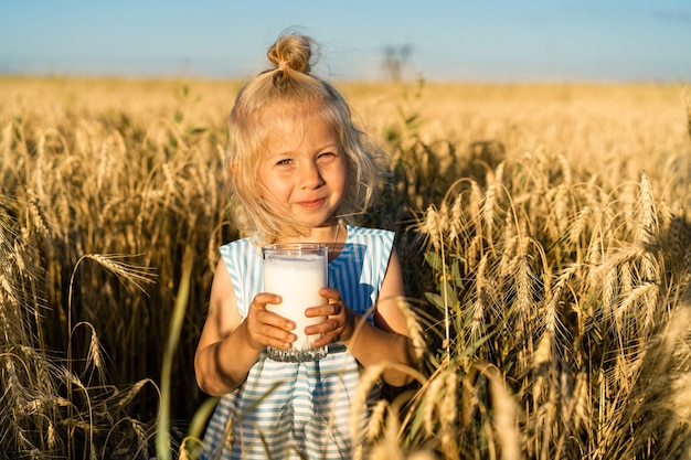 Mała blondynka na polu żyta uśmiecha się, szczęśliwe dziecko w uszach ze szklanką mleka