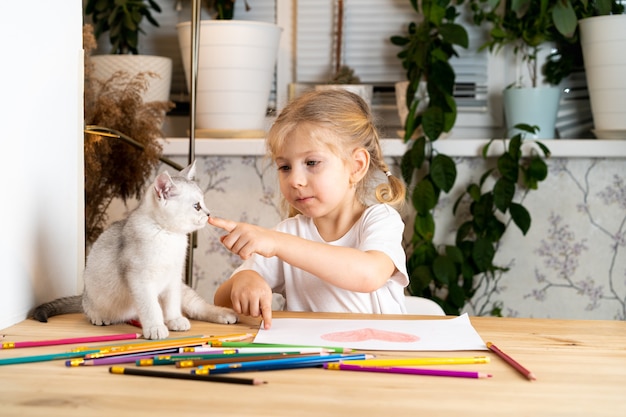 mała blondynka siedzi przy stole i uczy białego szkockiego kotka, kredek i kredki