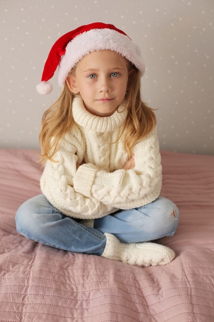 mała blondynka siedzi w domu na łóżku w zimowym swetrze i czapce Mikołaja