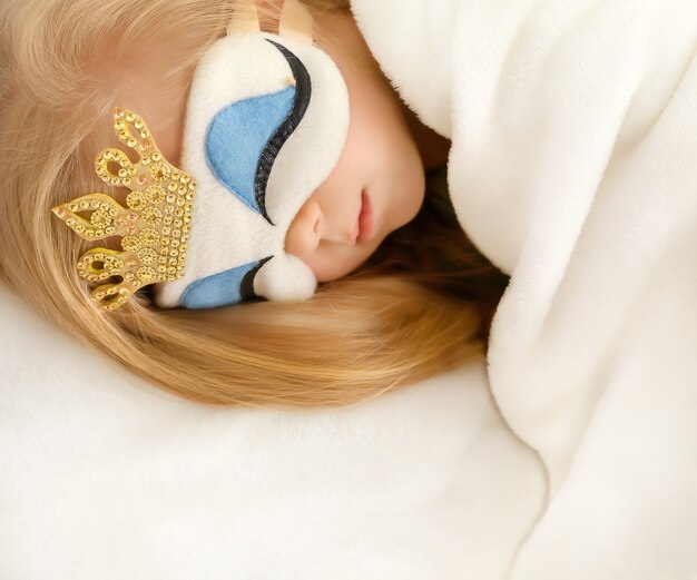 Zdjęcie mała blondynka śpi w łóżku pod białym kocem w masce śpiącej księżniczki