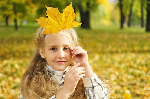 Mała blondynka trzymająca w dłoniach jesienny liść i patrząca w kamerę Zdjęcie poziome