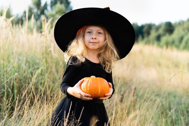 Mała blondynka w czarnym kostiumie i kapeluszu wiedźmy, Halloween z dynią w dłoniach, wesołe dziecko.