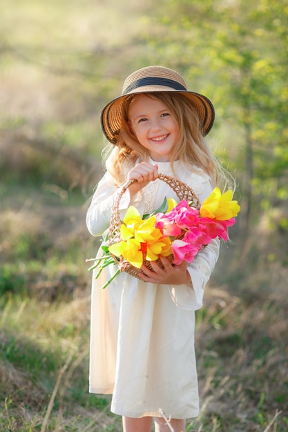 mała blondynka w słomkowym kapeluszu trzyma w naturze kosz wiosennych kwiatów
