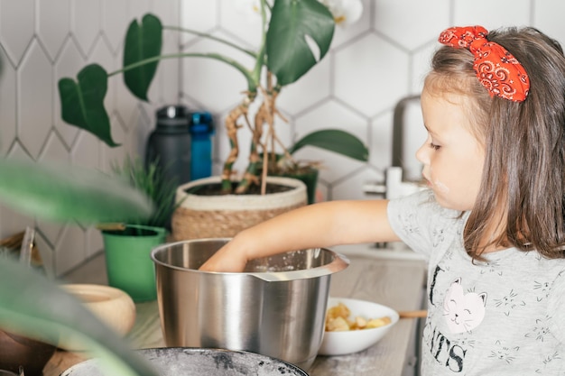 Mała ciemnowłosa dziewczynka 3 lata w czerwonej opasce piecze szarlotkę w kuchni. Dziecko dodaje jabłka do ciasta. Gotowanie dla dzieci