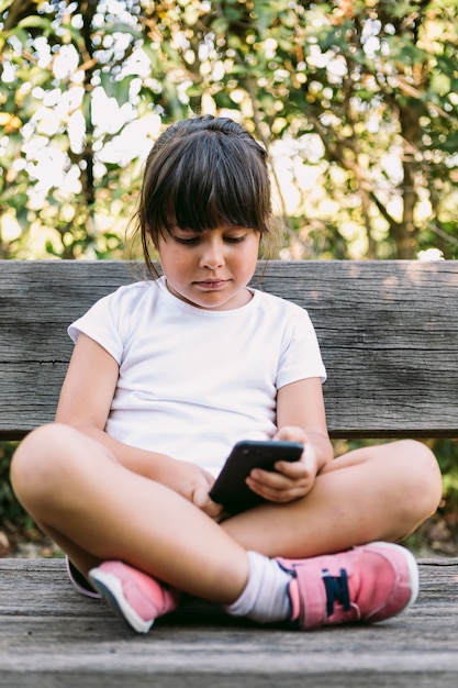 Mała czarnowłosa dziewczynka, ubrana w białą koszulkę, siedzi na ławce w parku, patrzy na telefon komórkowy, uśmiecha się.