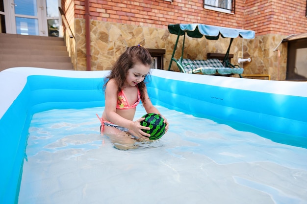 Mała dziewczynka bawi się pływającą piłką podczas zabawy w nadmuchiwanym basenie z wodą na domowym podwórku w słoneczny dzień
