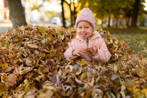 Mała dziewczynka bawi się w stercie jesiennych liści Mała dziewczynka bawi się w stercie jesiennych liści dziewczyna pokazuje język