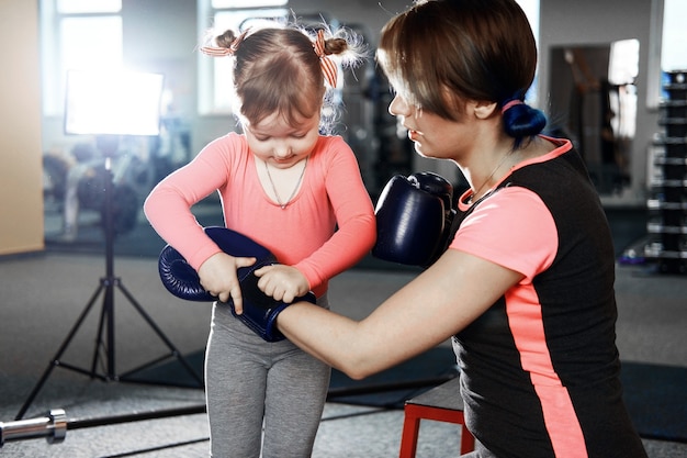 Mała dziewczynka ćwiczy boks, dziewczynka zakłada rękawice bokserskie, aby mama, mama i córka szykowały się do walki