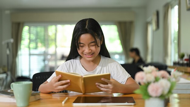 Mała dziewczynka czyta książkę przy drewnianym biurku. Koncepcja studia w domu.