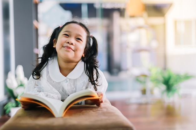 Zdjęcie mała dziewczynka czyta książkę w swoim pokoju na kanapie z pięknym uśmiechem.