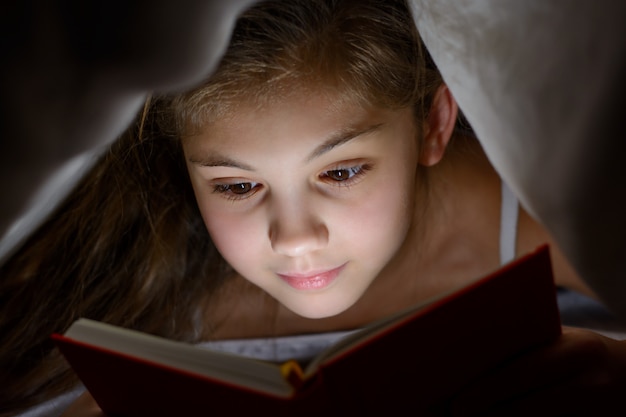 Mała dziewczynka czyta książkę z latarką pod kocem w łóżku