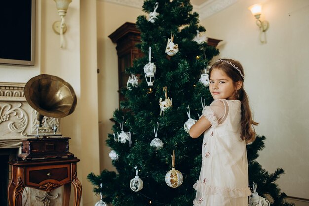 Mała dziewczynka dekoruje zabawki Choinka świąteczna w salonie w przytulnym domu Rodzinny moment atmosferyczny Wesołych Świąt Szczęśliwego Nowego Roku koncepcja Skopiuj miejsce na tekst