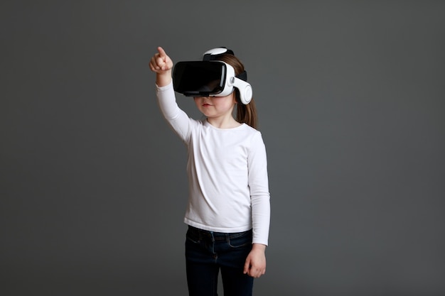 Mała dziewczynka doświadczająca okularów wirtualnej rzeczywistości w białej koszuli z długim rękawem na szarej powierzchni.