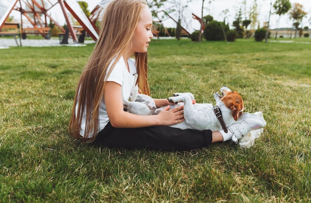 Mała dziewczynka drapie się po brzuchu swojego psa rasy Jack Russell Terrier Szczęśliwy pies leży na zielonej trawie w parku zwierzak