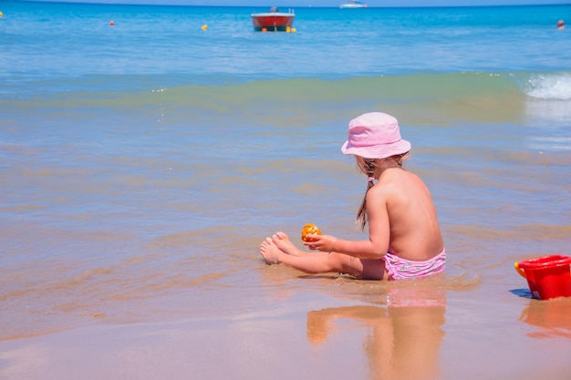 Mała Dziewczynka Gra Na Plaży Z Czerwonym Wiadrem I łopatą. Małe Dziecko Maluch Ssanie W Wodzie Sam Z Kapelusza W Słoneczny Letni Dzień. Dzieci Bawiące Się Zabawkami Na Plaży Na Tropikalnej Plaży. Skopiuj Miejsce