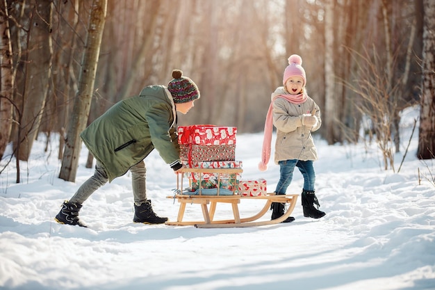 Mała dziewczynka i chłopiec niosą sanki z prezentami świątecznymi na zimowym tle