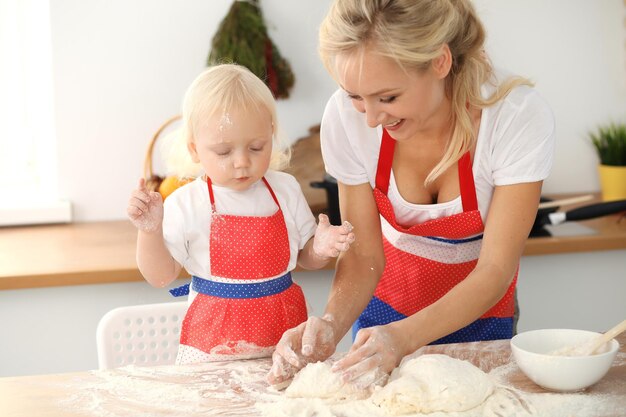 Mała dziewczynka i jej blond mama w czerwonych fartuchach bawią się i śmieją podczas wyrabiania ciasta w kuchni. Domowe ciasto na chleb, pizzę lub upiecz ciasteczka. Rodzinna zabawa i koncepcja gotowania.