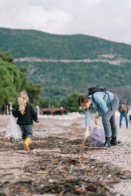 Mała dziewczynka idzie w pobliżu matki z torbą pomagającą jej zbierać śmieci na plaży