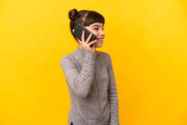 Mała dziewczynka kaukaski na białym tle na żółtej ścianie, prowadząc z kimś rozmowę z telefonem komórkowym
