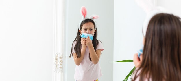 Mała Dziewczynka Kaukaski Z Uszami Królika I Maską Medyczną Na Twarzy Na Białym Tle. Koncepcja Wielkanocy. Ochrona Przed Koronawirusem