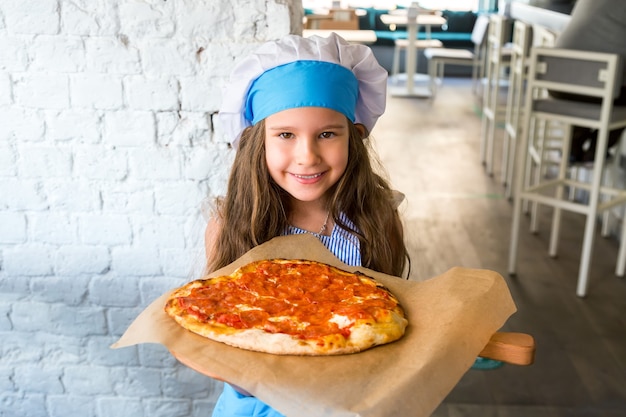 Mała dziewczynka kucharz uśmiechając się w pobliżu pizzy pepperoni w kształcie serca po przygotowaniu.