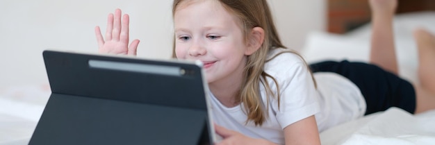 Mała dziewczynka leżąca na łóżku i machająca ręką na ekranie tabletu