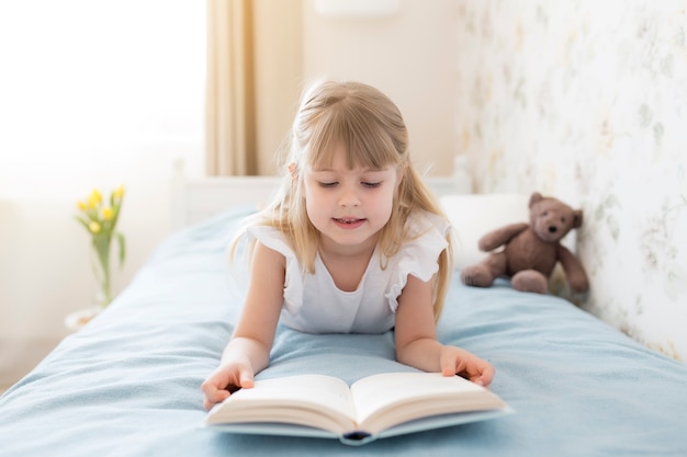 Mała dziewczynka leży na łóżku w stylowej sypialni i czyta niebieską książkę, odrabiając lekcje. Edukacja, koncepcja nauczania w domu. Żółte tulipany w wazonie przy łóżku