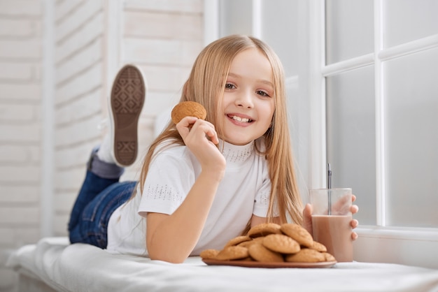 Mała dziewczynka leży na parapecie z ciasteczkami i mlekiem czekoladowym