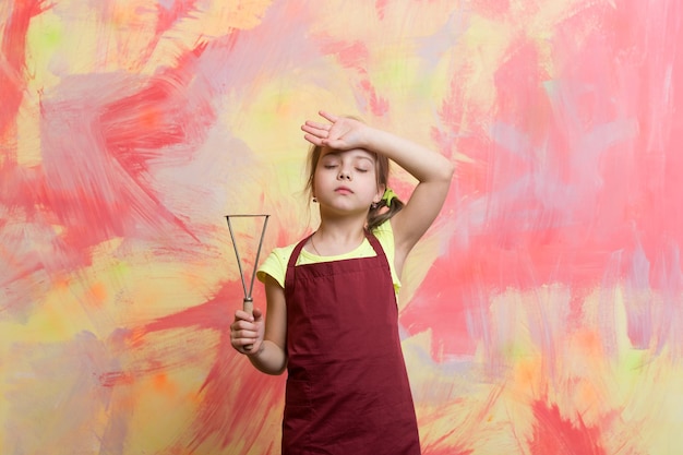 Mała dziewczynka lub słodkie dziecko ze zmęczoną twarzą w czerwonym fartuchu szefa kuchni lub kucharza trzymającego narzędzie do gotowania lub sprzęt kuchenny do puree na kolorowym abstrakcyjnym tle