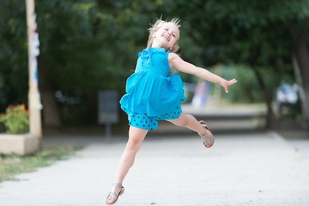 Mała dziewczynka lub słodkie szczęśliwe dziecko z uroczą uśmiechniętą twarzą i kokardą w blond włosach w niebieskiej sukience skaczące latem na zewnątrz na niewyraźne tło