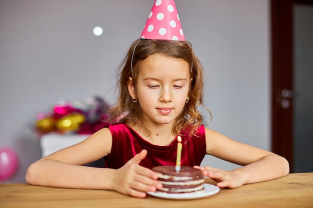 Mała Dziewczynka Ma Na Sobie Urodzinową Czapkę I życzy Sobie, Patrząc Na Tort Urodzinowy Ze świecącymi świeczkami Na Przyjęcie Z Okazji Uroczystości