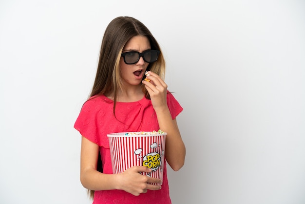 Mała dziewczynka na białym tle w okularach 3d i trzymająca duże wiadro popcornów, patrząc w bok