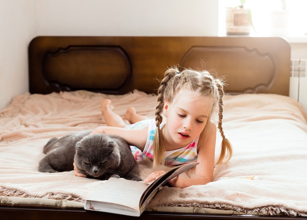 Mała Dziewczynka O Blond Włosach Leży Na łóżku W Domu Z Kotem Czytającym Książkę. Zostań W Domu. Edukacja Domowa Dla Dzieci.