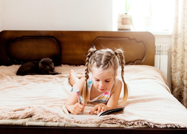 Mała dziewczynka o blond włosach leży na łóżku w domu z kotem czytającym książkę. Zostań w domu. Edukacja domowa dla dzieci.