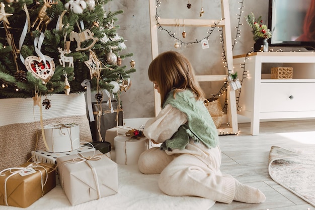 Mała dziewczynka o blond włosach siedzi na podłodze w pobliżu choinki z prezentami.