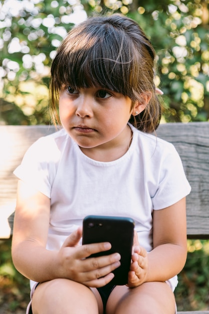 Mała dziewczynka o czarnych włosach, ubrana w białą koszulkę, siedząca na ławce w parku, patrząca na telefon komórkowy, poważnym gestem.