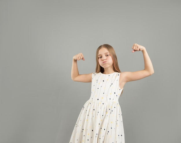 Mała dziewczynka pokazuje swoją siłę. Piękna młoda dziewczyna demonstrując siłę swoich ramion pozuje w lekkiej lekkiej sukience w studio na szarym tle.