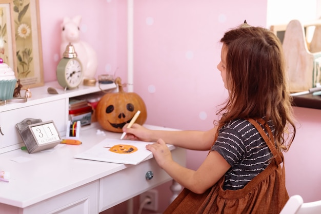 Mała dziewczynka rysuje dynię i przygotowuje się do świętowania Halloween w domu